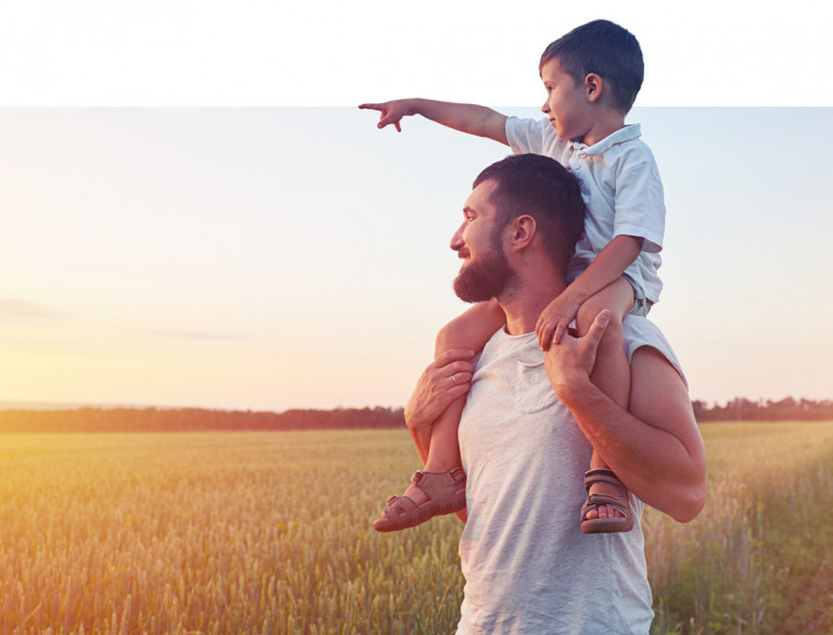 Small boy sitting on father’s shoulders in the field during sun